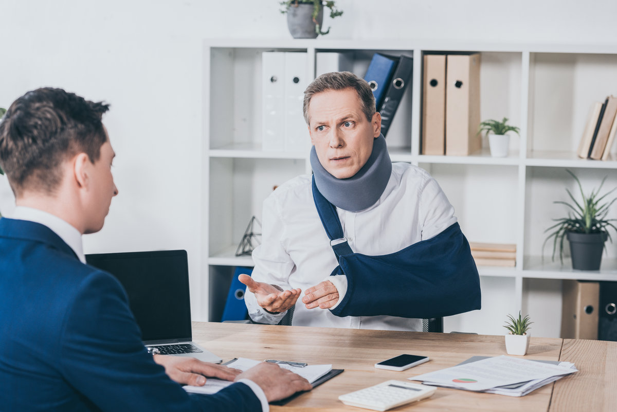 middle agaed worker in neck brace with broken arm sitting at table and talking to businessman in blue jacket in office, compensation concept