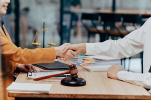 Legal Agreement Handshake: An agreement is sealed with a firm handshake between two individuals at a wooden table, with the gavel and scales of justice laid out in the background.