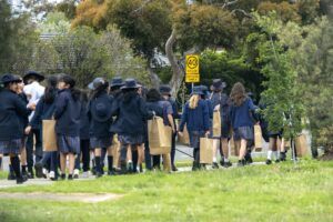 A yellow 40 km/h school zone sign on a roadside in Australia, with a group of uniformed school students walking along a suburban footpath in the foreground.